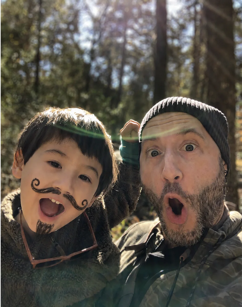 Blake and his son, one of them wearing a drawn-on mustache, both mid-yell in the woods